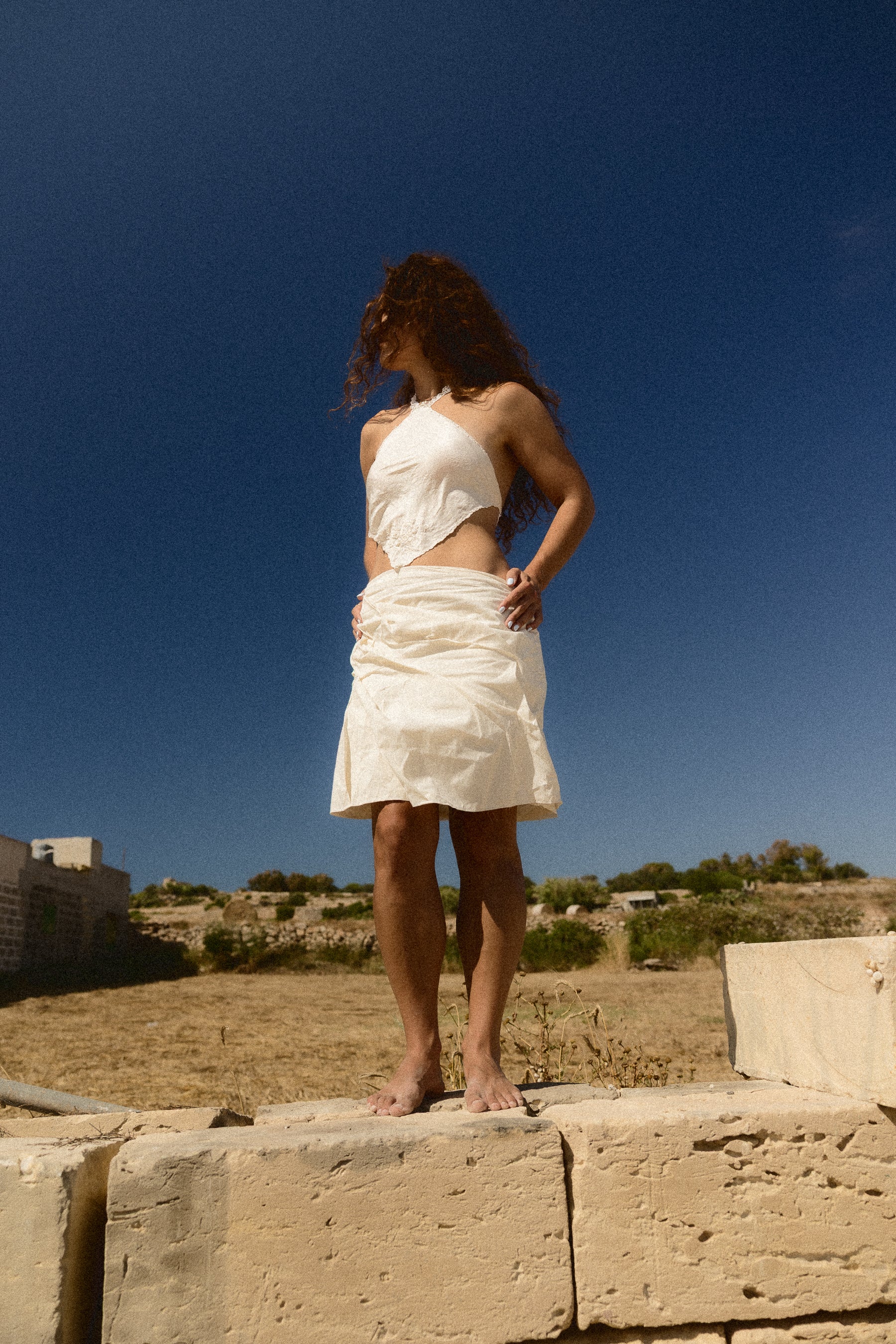 A woman stands outdoors on stone blocks under a deep blue sky, wearing a white sleeveless top and a matching white wrap skirt.