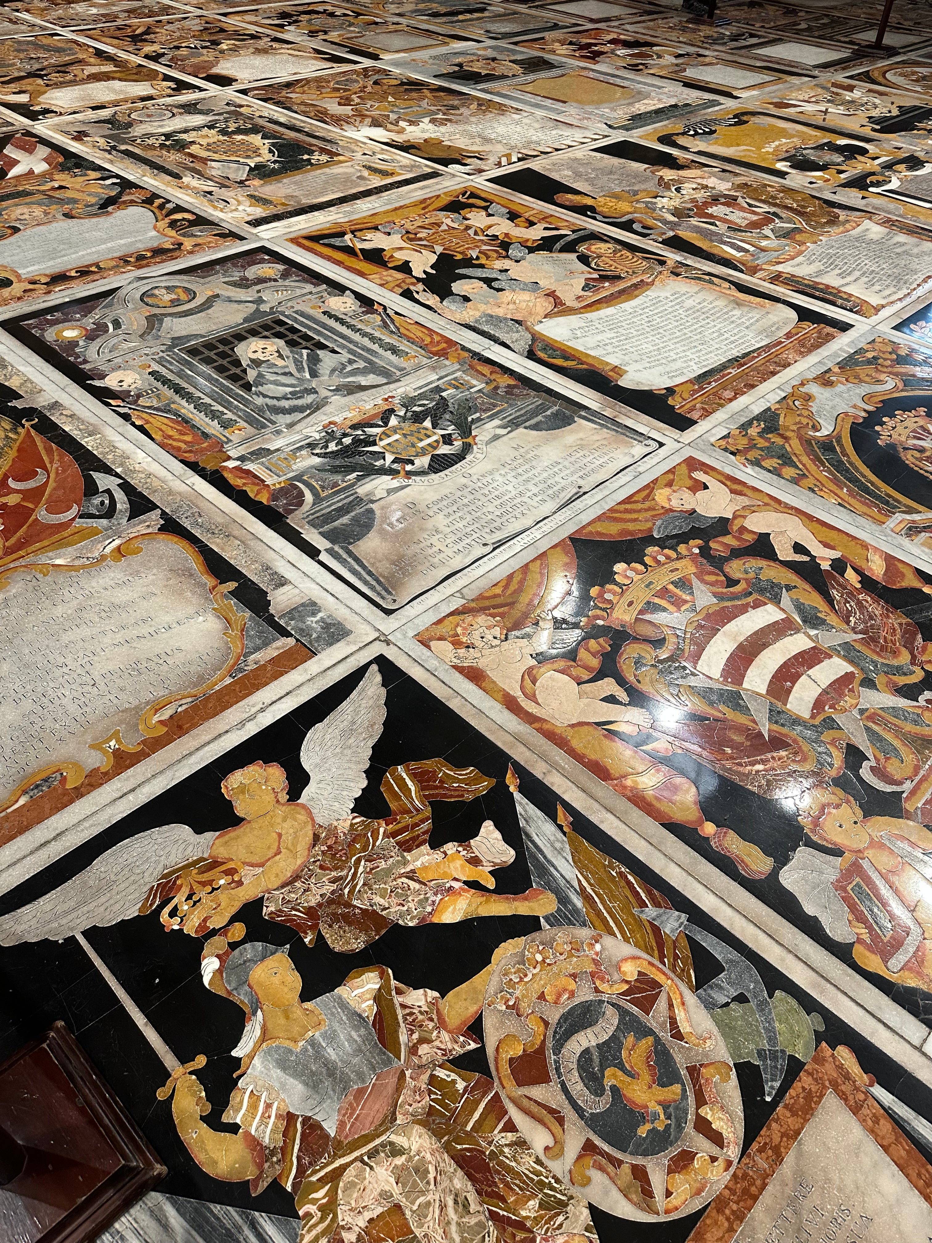 A close-up view of a highly detailed inlaid floor featuring religious and classical mosaic panels with cherubs, angels, script, and ornate decorative motifs in black, gold, beige, and red tones.