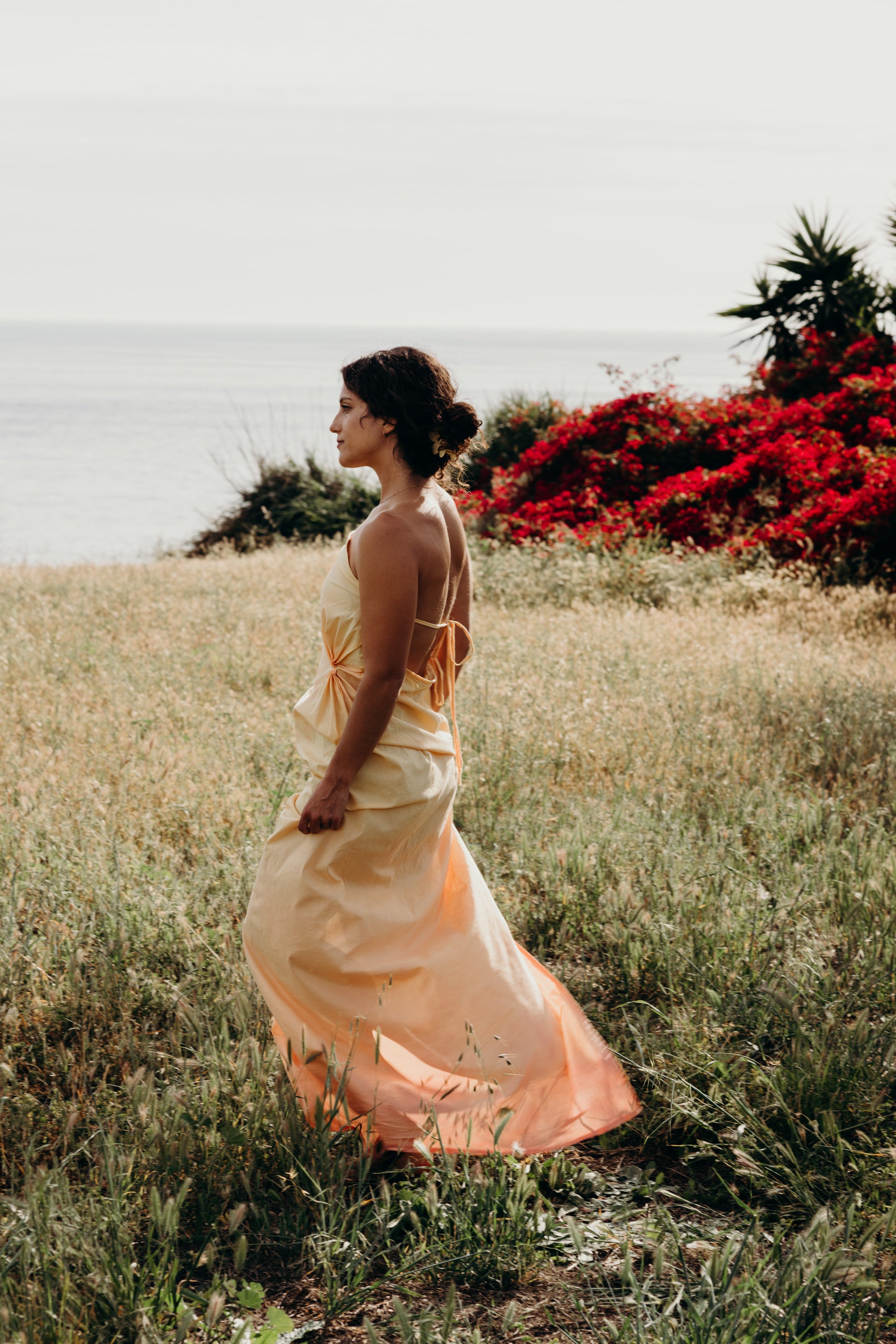 Woman in a flowing dress standing in a field with red flowers and a clear sky.