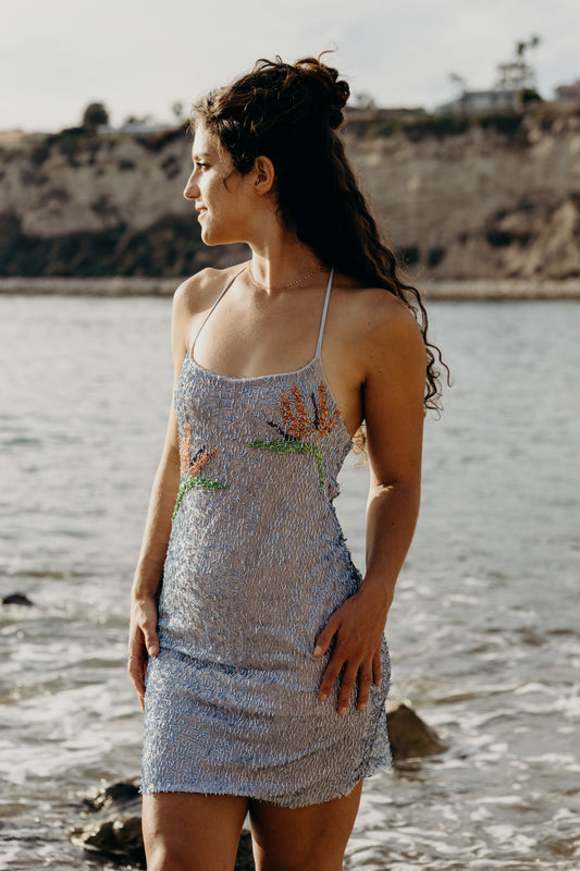 Woman in a sequined dress standing by a body of water with a scenic background.