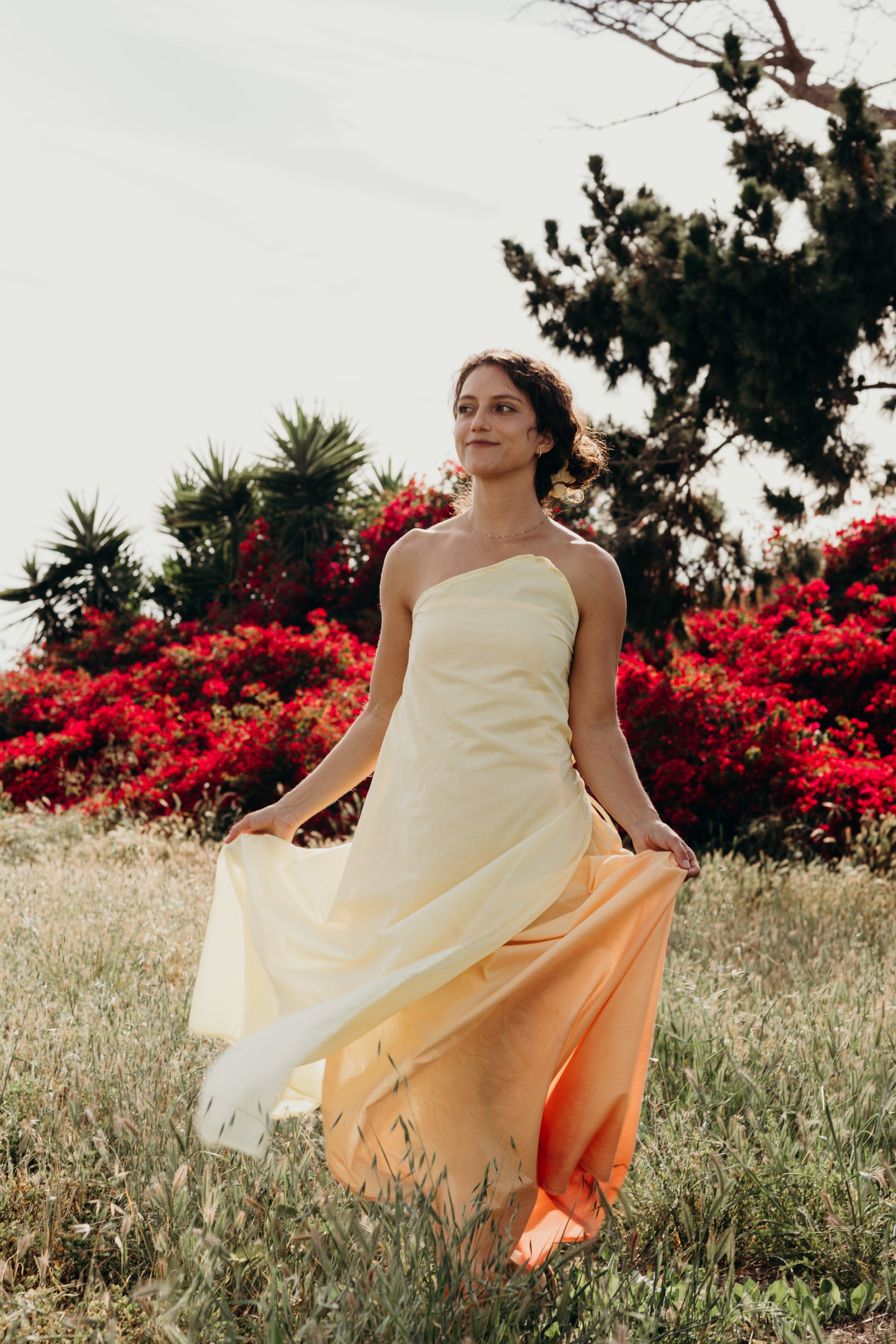 Woman in a strapless dress with a gradient from white to orange standing in a field with red flowers and trees.