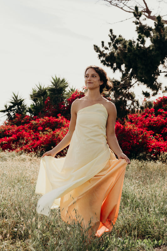 Woman in a strapless dress with a gradient from white to orange standing in a field with red flowers and trees.
