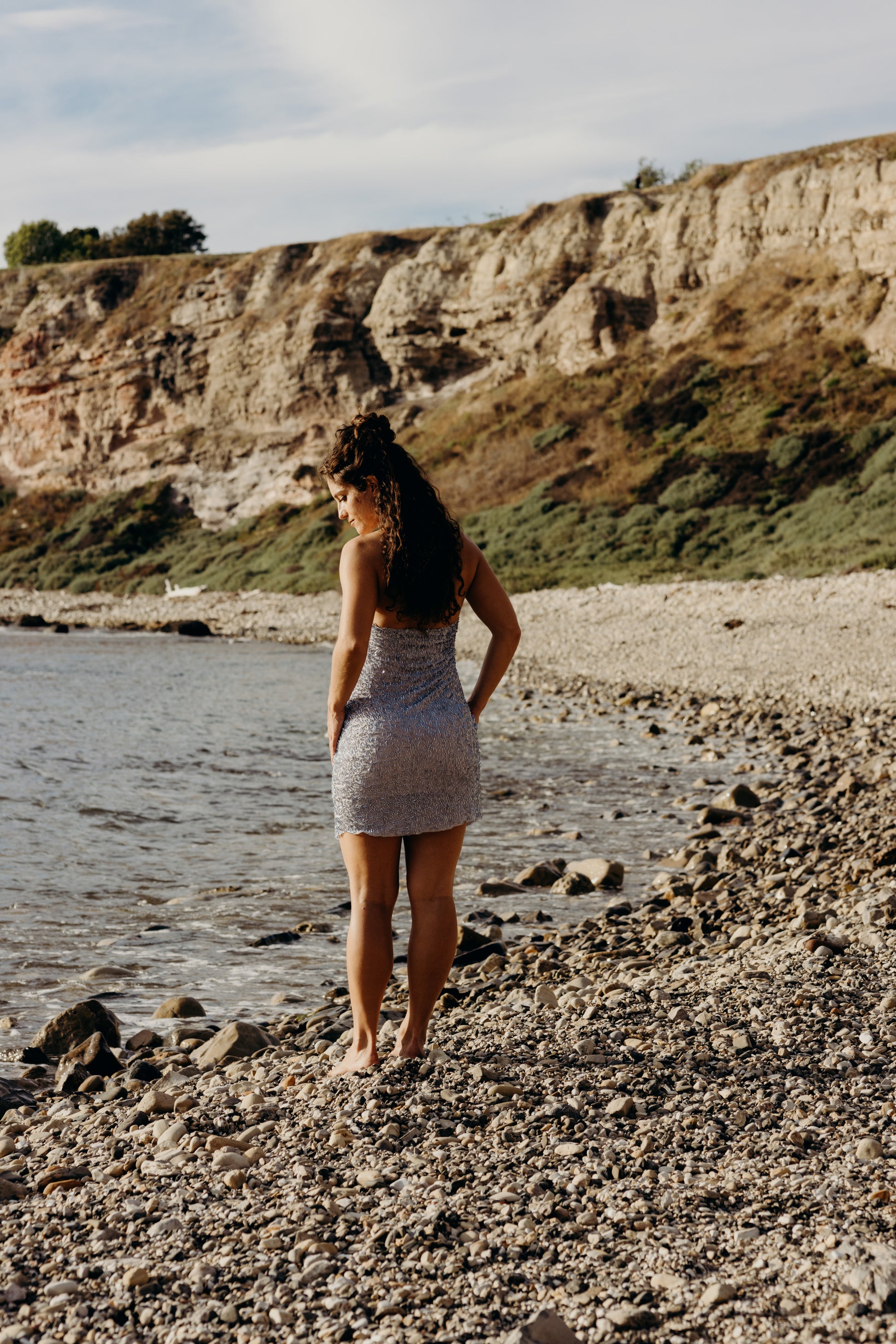 Woman in a silvery blue dress standing on a rocky beach with cliffs in the background