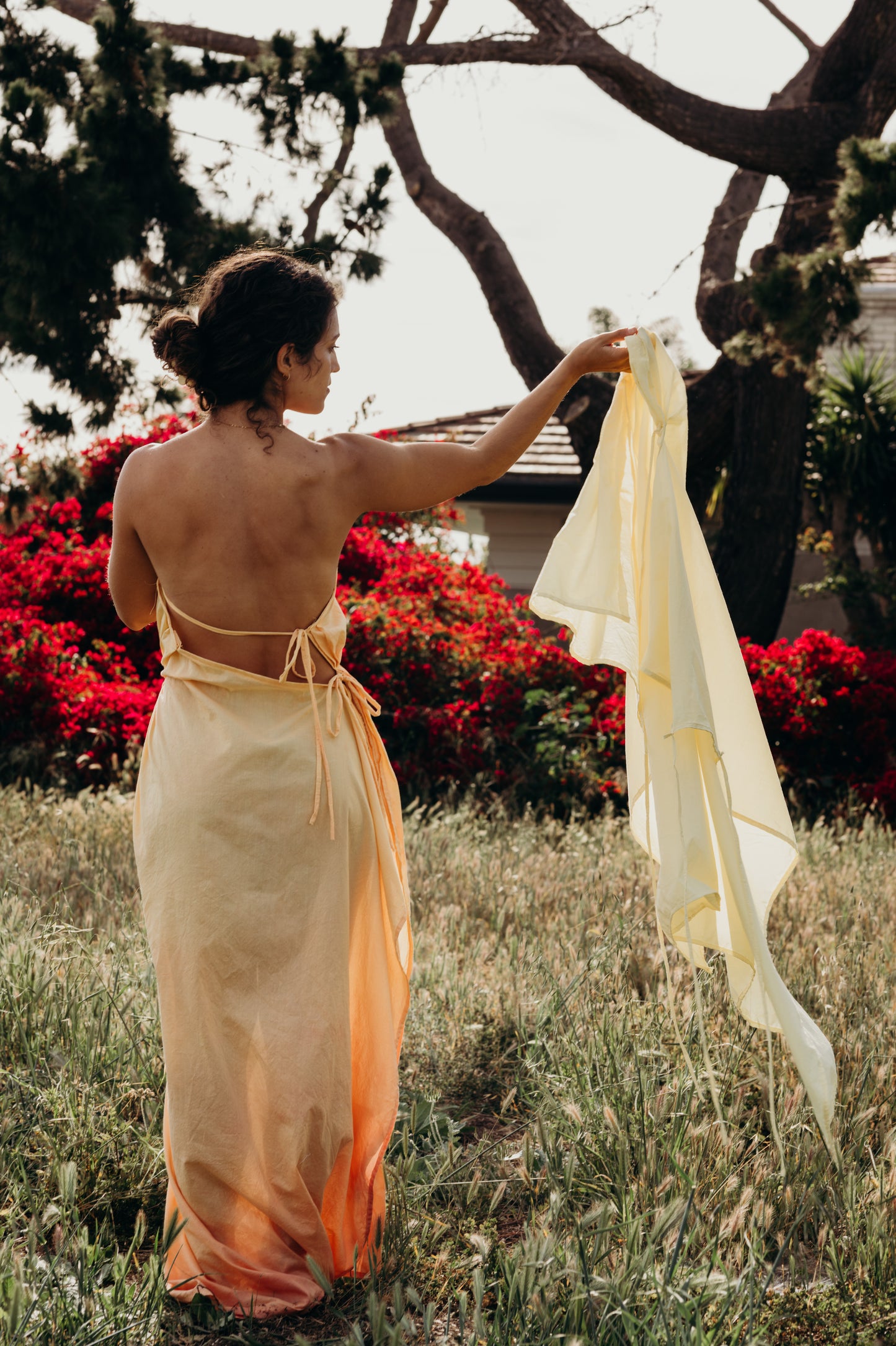 Woman in an orange dress holding a yellow dress in a garden with red flowers and trees.