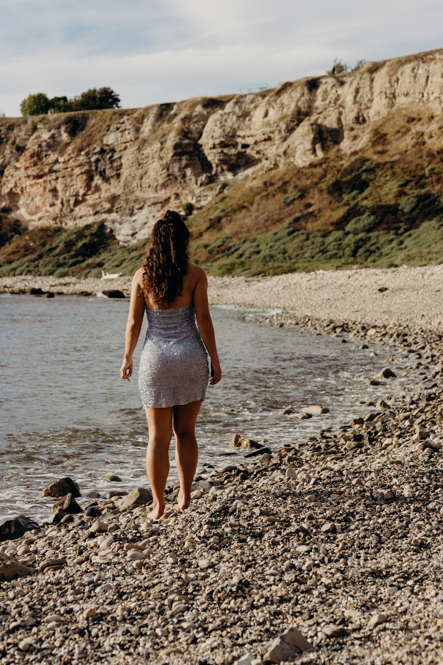 Woman in a silver blue dress standing on a rocky beach with cliffs in the background